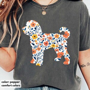 May include: A charcoal gray short-sleeve t-shirt featuring a large graphic of a dog silhouette filled with a floral pattern of orange and yellow flowers with blue leaves. The shirt is labeled as pepper color and comfort colors brand.