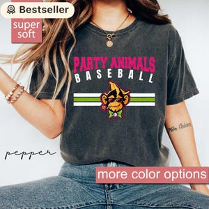 May include: Dark gray t-shirt with the text "PARTY ANIMALS BASEBALL" in pink and white. A cartoon monkey wearing sunglasses and a bow tie is in the center. The shirt has a green and white stripe design. The words "super soft" and "Bestseller" are also visible.