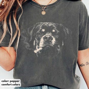 Könnte beinhalten: Ein anthrazitfarbenes Kurzarm-T-Shirt mit einem Grafikdruck eines Rottweilers, der eine Zigarette im Maul hält. Das T-Shirt hat einen Rundhalsausschnitt und eine verwaschene Vintage-Optik. Die Hundeillustration ist in Weiß gehalten.