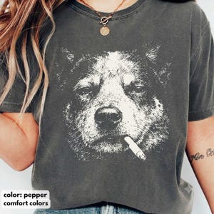 以下が含まれることがあります： チャコールグレーの半袖Tシャツで、犬の顔の大きく白い高コントラストなグラフィックプリントが施されています。犬は口にタバコをくわえた姿で描かれています。Tシャツはクルーネックで、ウォッシュ加工されたヴィンテージ風の生地の質感が特徴です。