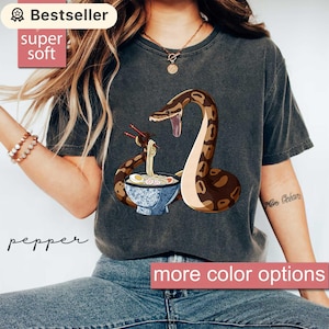 May include: Dark gray t-shirt featuring a brown and tan snake with a bowl of ramen. The snake is holding chopsticks with noodles. The text "Bestseller" and "super soft" are visible on the top left.