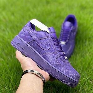 Könnte beinhalten: Violette Air Force 1 Sneaker mit Schlangenlederstruktur. Die Sneaker haben eine durchgehende violette Sohle, Schnürsenkel und einen Nike-Swoosh. Ein zweiter Sneaker ist im Hintergrund zu sehen. Die Sneaker werden vor einem grünen, grasbewachsenen Hintergrund gehalten.