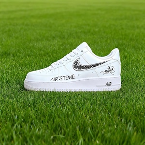 Pode incluir: Tênis de couro branco com design de swoosh preto e uma figura de desenho animado na lateral. O texto "AIR'STEWE" é impresso na lateral do tênis, e a palavra "AIR" é impressa na sola. O tênis está sobre grama verde.