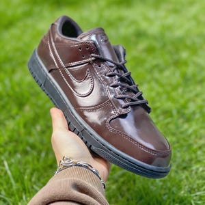 Può includere: Una sneaker bassa marrone e nera con finitura lucida. La scarpa presenta una suola nera, lacci e un logo Nike swoosh sul lato. La sneaker è tenuta su uno sfondo di erba verde.