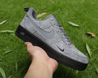 May include: Gray suede Air Force 1 sneaker with a black sole. The shoe features a gray Nike swoosh and the word "AIR" in white on the side. The sneaker is held against a background of green grass.