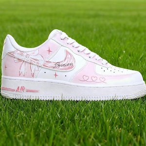 May include: White and pink Air Force 1 trainer with a princess theme. The shoe features a pink swoosh, bow, and the word "Princess" in script. Pink stars and hearts decorate the side. The word "AIR" is printed in pink on the sole.