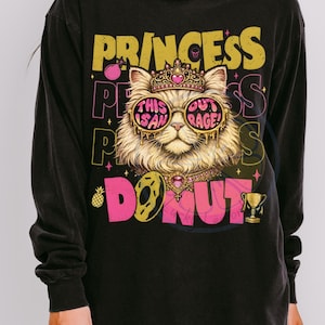 May include: Black long-sleeve shirt with a graphic of a cat wearing sunglasses, a crown, and a necklace. The text reads "PRINCESS DONUT" with the phrases "THIS IS AN OUTRAGE!" on the sunglasses.