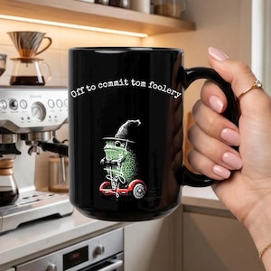 May include: Black ceramic mug with the phrase "Off to commit tom foolery" in white script. The mug features a green frog wearing a witch hat and riding a red scooter. The mug has a black handle.