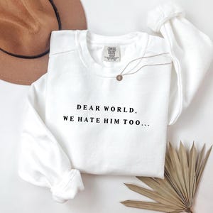 Peut inclure: Un sweat-shirt blanc avec le texte "DEAR WORLD, WE HATE HIM TOO..." en noir. Le sweat-shirt a des manches longues et un col rond. Un collier doré et un chapeau marron sont également visibles.