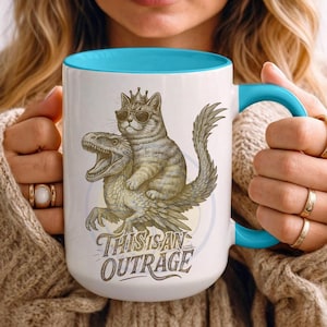 以下が含まれることがあります： 青い内側とハンドルが付いた白いセラミックマグ。サングラスと王冠をつけた猫が恐竜に乗っているユーモラスなイラストが特徴です。「THIS IS AN OUTRAGE」というテキストがイラストの下にあります。