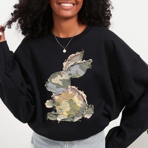 Può includere: Felpa nera con un design a coniglio patchwork. Il coniglio è realizzato in tessuto mimetico nei toni del verde, marrone e beige. La felpa ha un girocollo e maniche lunghe, ideale per l'abbigliamento casual.
