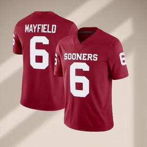 May include: Two crimson football jerseys. One jersey displays "MAYFIELD" and the number "6" in white. The other jersey shows "SOONERS" and the number "6" in white. Both jerseys have a v-neck design.