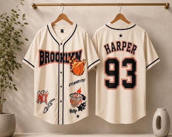 Może przedstawiać: Kremowa koszulka baseballowa zapinana na guziki z czarno-pomarańczowymi grafikami. Z przodu widnieje napis 'BROOKLYN', płonąca piłka do koszykówki, 'NY', 'CHAMPION' oraz kosz. Z tyłu znajduje się napis 'HARPER' i numer '93' w kolorze czarnym i pomarańczowym.