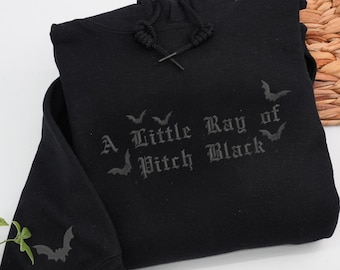 May include: Black hooded sweatshirt with the phrase "A Little Ray of Pitch Black" and bat graphics. The sweatshirt has a drawstring hood and a bat graphic on the sleeve. The garment is folded on a white surface.