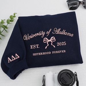 May include: A navy blue sweatshirt with pink embroidered text reading "University of Alabama", a bow, "EST. 2025", and "SISTERHOOD FOREVER". The sleeve has "ΑΔΔ". Sunglasses and a camera are visible.