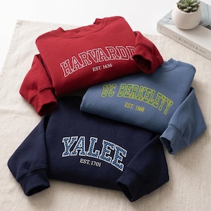Könnte beinhalten: Drei Rundhals-Sweatshirts in Rot, Blau und Marineblau. Das rote Sweatshirt zeigt den weißen Schriftzug HARVARD EST. 1636. Das blaue Sweatshirt hat den gelben Schriftzug UC BERKELEYY EST. 1868. Das marineblaue Sweatshirt hat den karierten Schriftzug YALEE EST. 1701.