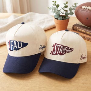 May include: Two baseball caps with cream-coloured crowns and navy blue brims. One cap features "FAU" in a pennant design, the other "STANFORD". Both have the name "Sophia" embroidered on the side. A football and books are in the background.