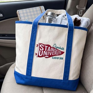 Può includere: Borsa tote in tela color crema con dettagli, manici e base blu. La borsa presenta il testo "Standford University" in rosso e bianco, con "Class of 2026" sotto. Contiene un quaderno, una bottiglia d'acqua e una felpa con cappuccio grigia.