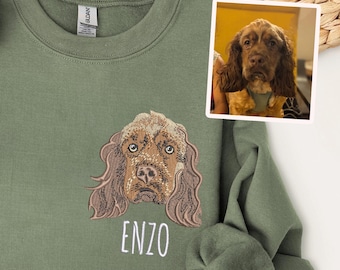 May include: An olive green crewneck sweatshirt featuring a detailed embroidered portrait of a brown spaniel dog with long ears. Below the dog, the name ENZO is embroidered in white capital letters. A small photo of the dog is attached to the top right.