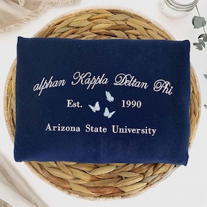May include: A navy blue sweatshirt with white embroidered text reading "alphan Kappa Delta Phi Est. 1990 Arizona State University." The design includes three small butterflies. The sweatshirt is folded on a woven, circular placemat.