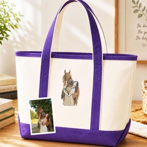 Puede incluir: Un bolso tote de lona color crema con ribetes y asas moradas. El bolso presenta un diseño bordado de una persona abrazando a un caballo. Una pequeña foto de una persona y un caballo está adherida al bolso.