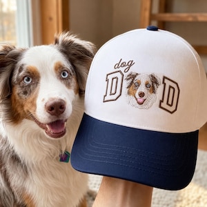 Puede incluir: Una gorra de béisbol blanca con visera azul oscuro y un botón azul oscuro en la parte superior. El frente presenta el texto bordado dog DAD, con la letra O reemplazada por una ilustración bordada de un perro marrón, blanco y de ojos azules.