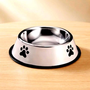 May include: A circular pet bowl made of polished stainless steel with a reflective silver finish. The exterior side of the bowl features two black paw print designs positioned opposite each other, and the base has a black rubberized rim.