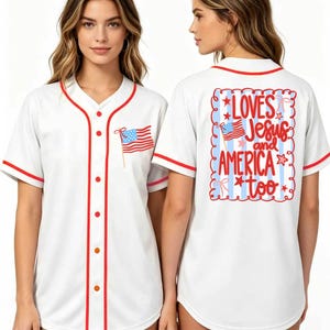 May include: A white baseball jersey with red trim, featuring a small American flag graphic on the front left chest. The back displays a blue and white striped rectangular design with the red text LOVES Jesus and AMERICA too.