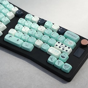 May include: A black keyboard with custom keycaps in shades of blue, white, and navy. The keycaps feature Cyrillic characters and decorative icons. The keyboard has a rose gold accent and a small touchpad.
