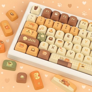 Może przedstawiać: Biała klawiatura mechaniczna z niestandardowymi nakładkami w odcieniach brązu, beżu i zieleni. Nakładki na klawisze mają wzory związane z jedzeniem i zwierzętami. Klawiatura znajduje się na brzoskwiniowej powierzchni z dekoracjami w kształcie serc i śladów łap.