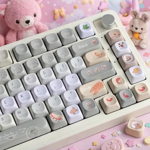 May include: A white mechanical keyboard with custom keycaps featuring various illustrations. The keycaps have a mix of gray, white, and beige colors, with images of a shrimp, a leaf, and other cute designs. The keyboard has a silver knob and a few gray function keys.