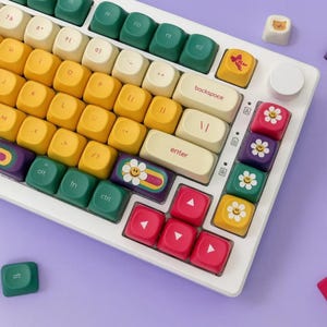 Puede incluir: Un teclado mecánico blanco con teclas de colores. Las teclas son amarillas, verdes, rojas y blancas, algunas decoradas con flores y otros diseños. El teclado tiene un marco blanco y una rueda de volumen redonda.
