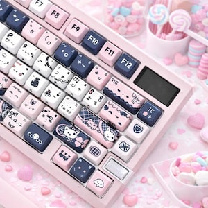 Puede incluir: Un teclado mecánico rosa con teclas blancas, azul marino y rosa. Las teclas presentan varias ilustraciones, incluyendo gatos, nubes y otros diseños bonitos. El teclado tiene una pequeña pantalla y está rodeado de caramelos rosas.