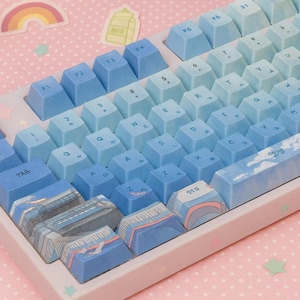 May include: A mechanical keyboard with blue gradient keycaps. The keycaps feature a range of blue hues, from light to dark, with white lettering. Some keycaps have illustrations of buildings and clouds. The keyboard sits on a pink surface.