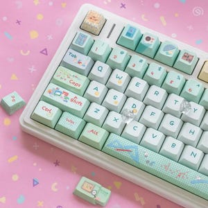 Könnte beinhalten: Eine weiße mechanische Tastatur mit pastellgrünen Tastenkappen. Die Tastenkappen zeigen verschiedene Symbole und Text, darunter "Tab", "Caps", "Shift" und "Win". Die Tastatur liegt auf einer rosa Oberfläche mit bunten geometrischen Formen.