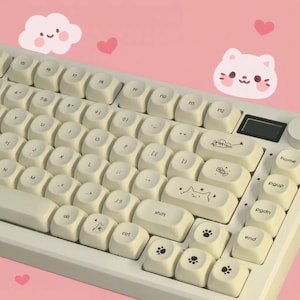 May include: A white mechanical keyboard with cream-colored keycaps. The keycaps feature cat-themed designs and paw prints. The background is pink with cartoon cloud and heart illustrations.