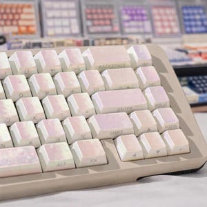 Puede incluir: Primer plano de un teclado mecánico con teclas iridiscentes. Las teclas tienen un acabado nacarado con tonos rosas y blancos. El teclado tiene un marco beige y una distribución de teclas estándar.