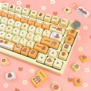 Op de afbeelding: Een aangepast mechanisch toetsenbord met crème en oranje keycaps. De keycaps hebben berenillustraties en andere schattige ontwerpen. Het toetsenbord heeft een zilveren knop en extra keycaps verspreid.
