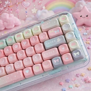 Może przedstawiać: Klawiatura w pastelowych kolorach z różowymi, niebieskimi i zielonymi klawiszami. Klawisze zdobią różne urocze wzory, takie jak serca, kokardy i chmury. Klawiatura jest w przezroczystej obudowie, z różowym tłem i elementami dekoracyjnymi.