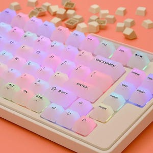 Può includere: Una tastiera meccanica bianca con tasti traslucidi dai colori pastello. I tasti sono illuminati con una sfumatura arcobaleno e alcuni tasti di ricambio sono sparsi sopra. La tastiera ha tasti con le etichette "ENTER", "SHIFT" e "BACKSPACE".