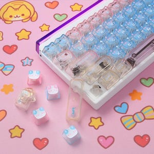 May include: A white mechanical keyboard with gradient pink and blue keycaps. Additional keycaps with a frog design are scattered on a pink surface decorated with hearts, stars, and a cat illustration.