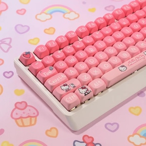 Könnte beinhalten: Eine pinke und weiße mechanische Tastatur mit pinkfarbenen Tastenkappen. Die Tastenkappen zeigen den Text "Hello Kitty" und verschiedene niedliche Illustrationen. Die Tastatur befindet sich auf einem pinkfarbenen Hintergrund mit Regenbogen- und Herzmustern.