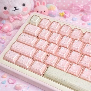 Può includere: Una tastiera rosa con tasti ricoperti di glitter. I tasti sono disposti in un layout QWERTY standard. La tastiera è circondata da peluche rosa e oggetti decorativi, creando un'estetica carina e giocosa.