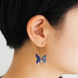 May include: A close-up of a gold-colored earring with a blue and white butterfly design. The butterfly has a red sunburst pattern and is attached to a gold hook. The earring is hanging from an ear.