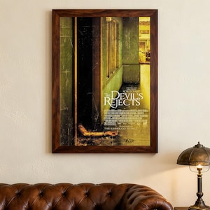 May include: A framed movie poster for "The Devil's Rejects" with a distressed look. The poster displays the film's title and credits, set against a dimly lit interior with a hand reaching out. The frame is a dark brown colour.