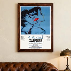 以下が含まれることがあります： 「アンディ・ウォーホル」と「Querelle」の文字が入った、2人の人物の青と赤のグラフィックが描かれた額入りのポスター。 ポスターは濃い茶色の木製フレームに入っており、クリーム色の壁に飾られています。