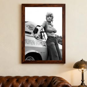 Puede incluir: Impresión en blanco y negro enmarcada que presenta a una mujer apoyada en un coche antiguo. El coche tiene el texto "Bulding Dodge" y "Romeo Palomides Race Cars". La mujer lleva un top corto y vaqueros.