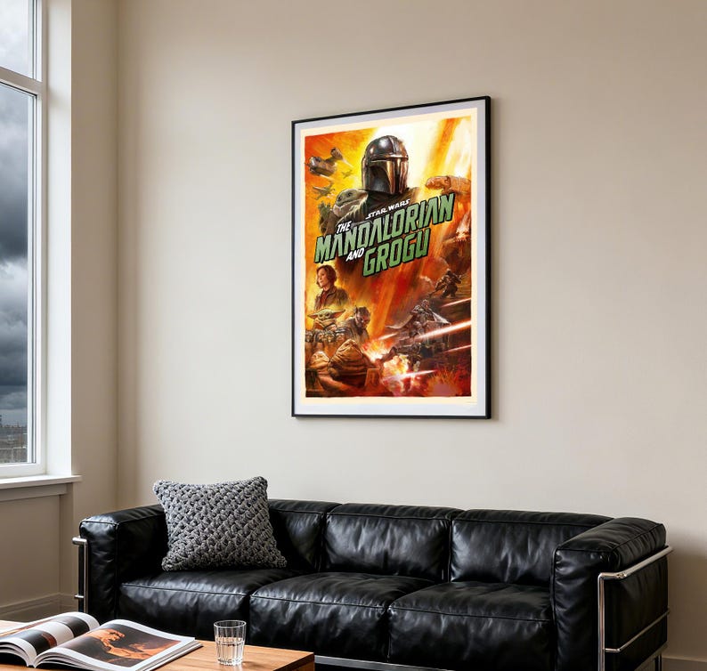 May include: A framed poster for Star Wars: The Mandalorian and Grogu. The artwork depicts the Mandalorian, Grogu, and other characters in a dynamic scene with a fiery background. The poster is displayed above a black leather sofa in a living room setting.