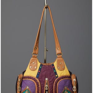 May include: A colorful patchwork handbag with brown leather straps and accents. The bag features a mix of purple, yellow, and teal panels, with decorative zippers and jewel-toned embellishments. The bag is displayed on a metal stand.