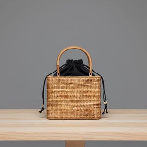 Puede incluir: Un bolso de mano cuadrado de ratán tejido con un asa redondeada. El bolso tiene una bolsa negra con cordón en el interior. El bolso está sobre una superficie de madera clara sobre un fondo gris.